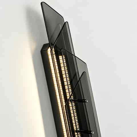 A close up of a modern wall lamp, showcasing layered smoke grey glass panels, a black metal frame, and built in LED lighting